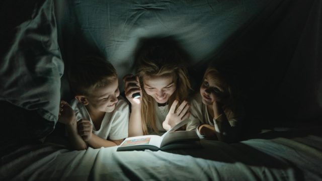 A women holding a torch, reading to a boy and girl while under a sheet.